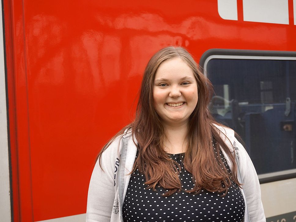 Eine junge Frau mit langen braunen Haaren steht lächelnd vor einem roten Zug.
