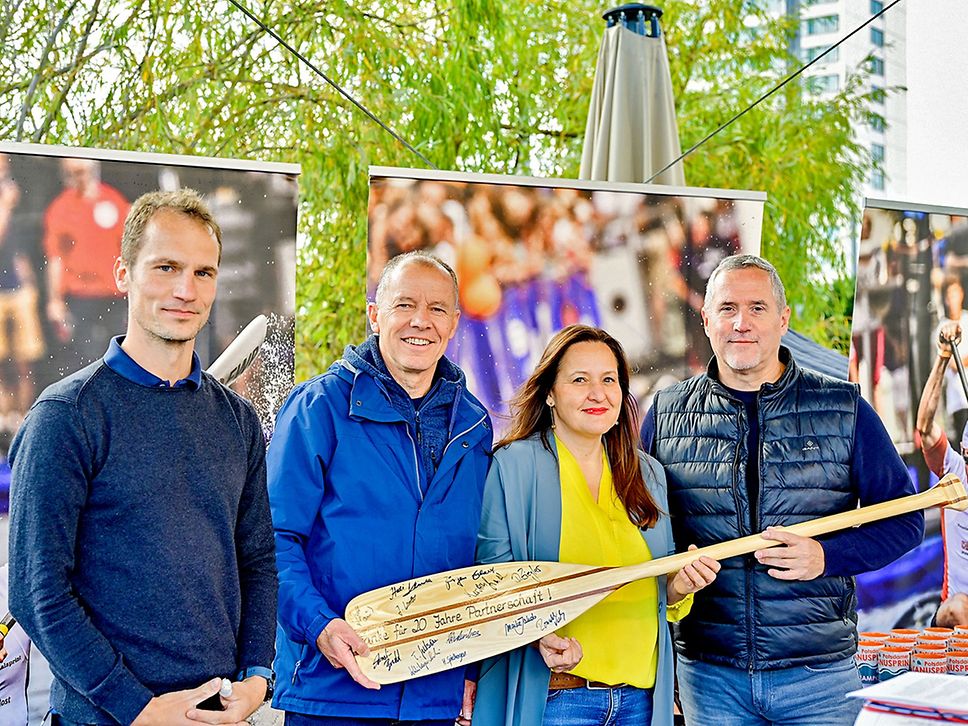Drei Männer und eine Frau lächeln in die Kamera, sie haben ein Paddel aus Holz in der Hand.