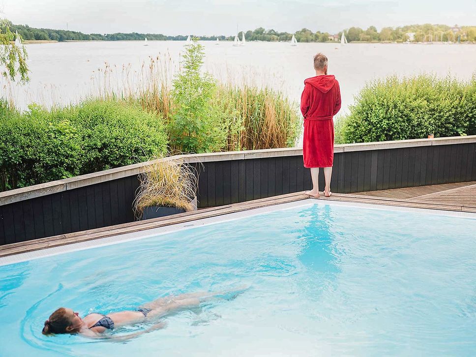 Ein Mann im roten Bademantel steht am Rand eines Pools und schaut über grüne Hecken auf einen See.
