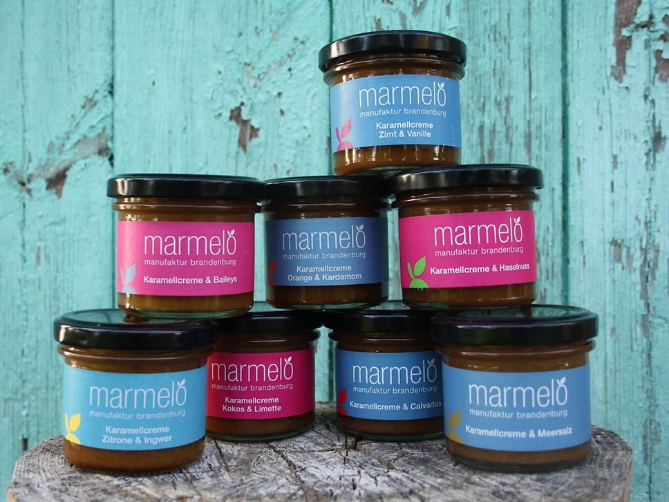 Acht kleine Gläschen mit Marmelade und Aufstrich stehen gestapelt vor einer blauen Holzwand.