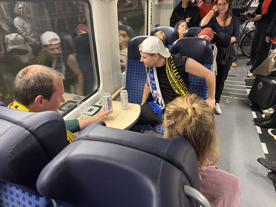 Zwei Männer mit Fanschals sitzen sich gegenüber und trinken Bier, daneben ist von hinte eine Frau zu sehen.