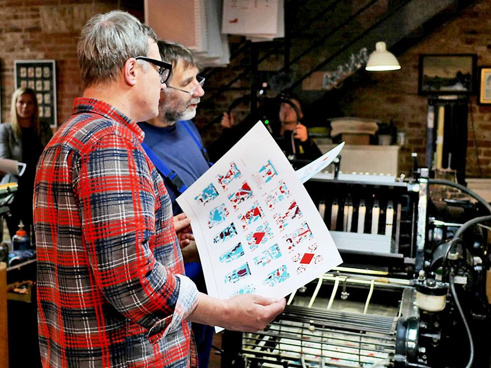 Zwei Männer halten Druckbögen in der Hand, darauf zu sehen sind Spielkarten.