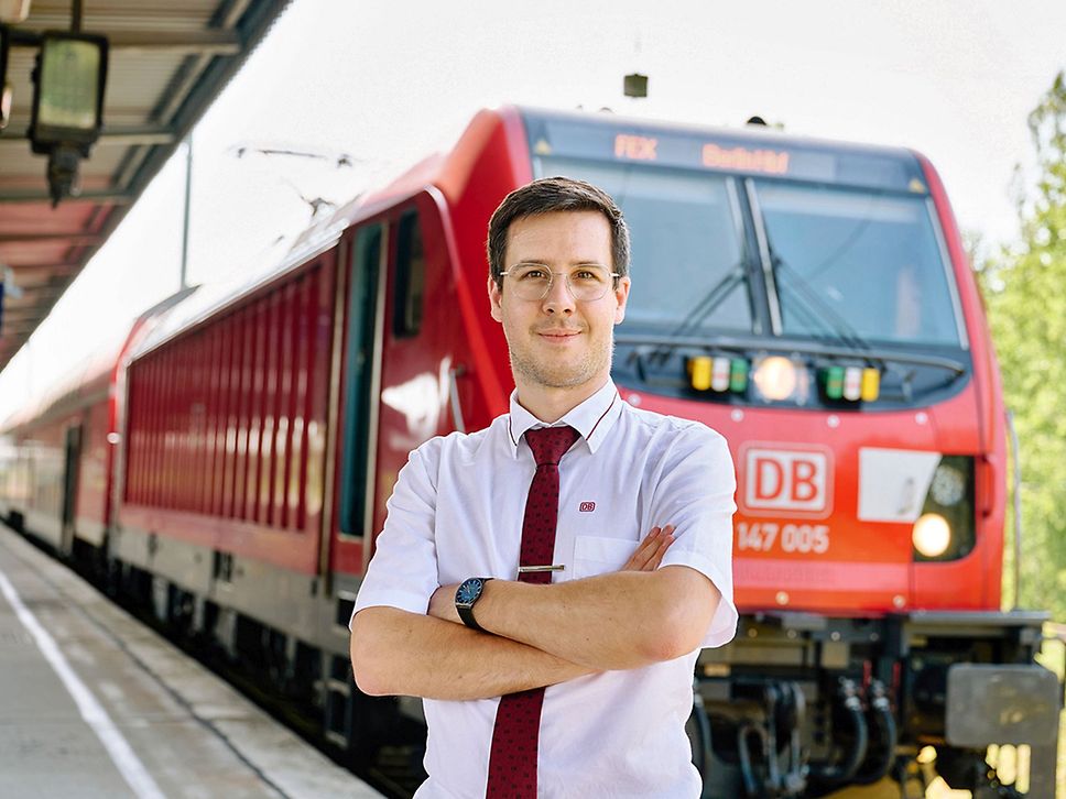 Ein junger Mann im weißen Hemd und mit roter Krawatte steht mit verschränkten Armen vor einer roten Lok.
