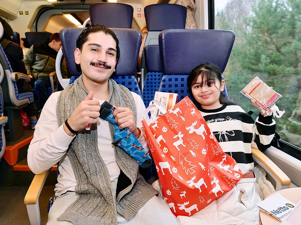 Ein Junge mit Schnurrbart sitzt im Zug neben einem Mädchen, sie packen eine rote Tüte mit Geschenken aus.