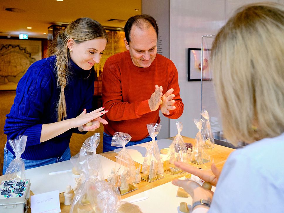 Eine Frau im blauen und ein Mann im roten Pulli rollen Kugeln aus Marzipan, eine weitere Frau ist von hinten zu sehen.