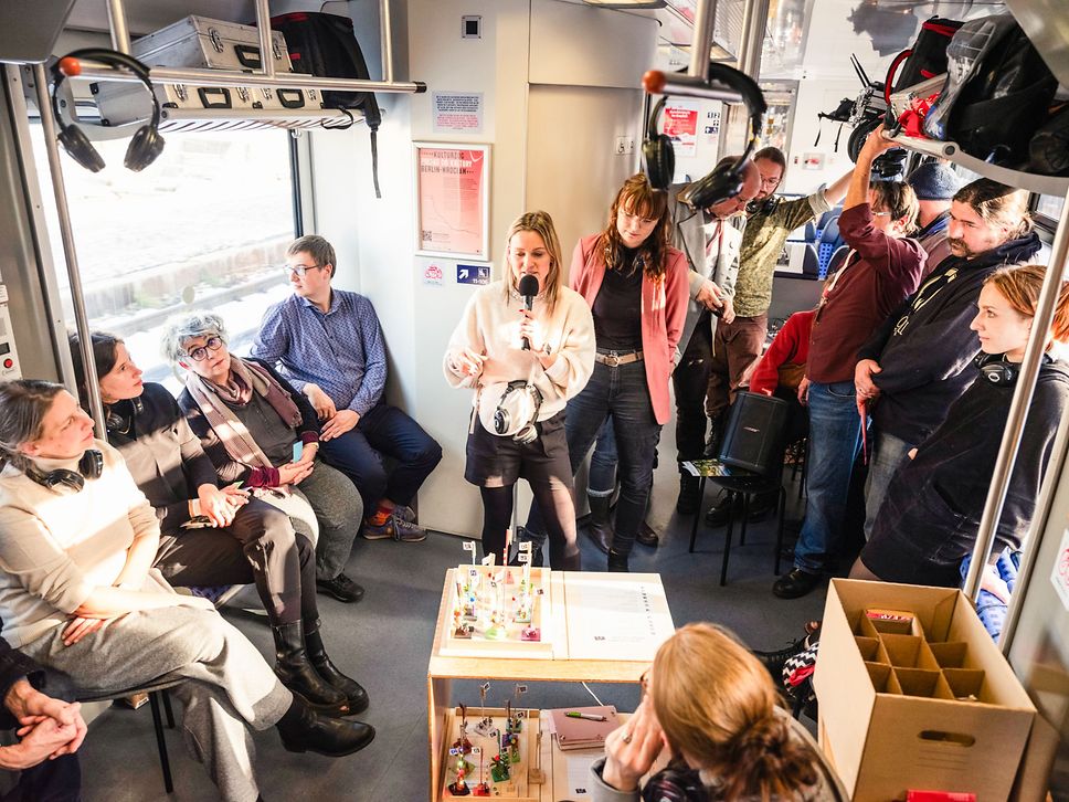 Eine Frau spricht in ein Mikrofon, sitzende und stehende Fahrgäste hören ihr zu.
