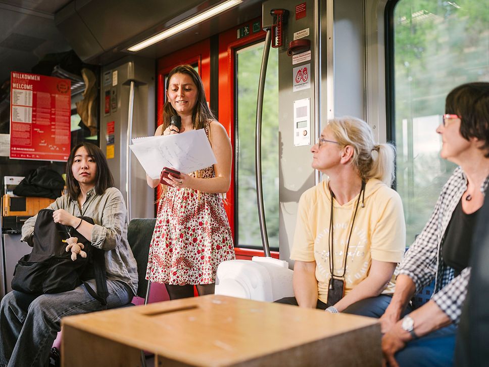 Eine Frau mit Mikrofon liest von einem Zettel ab, drei Frauen sitzen daneben und hören zu.