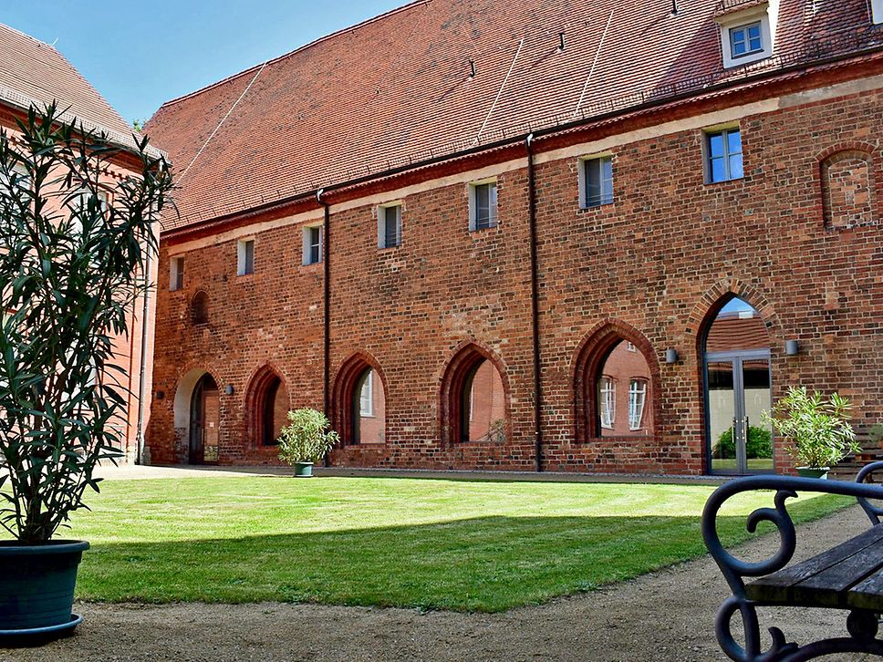 Blick in den Klosterhof, mittig grüne Wiese, rechts am Rand eine Bank, links eine Grünpflanze im Topf.