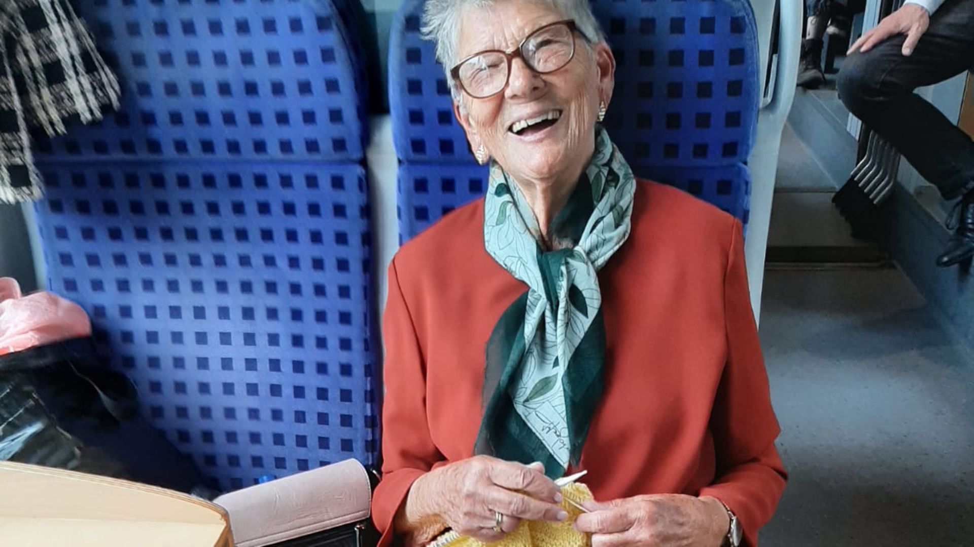 Eine gut gelaunte Frau strickt an einem Schal im Kulturzug