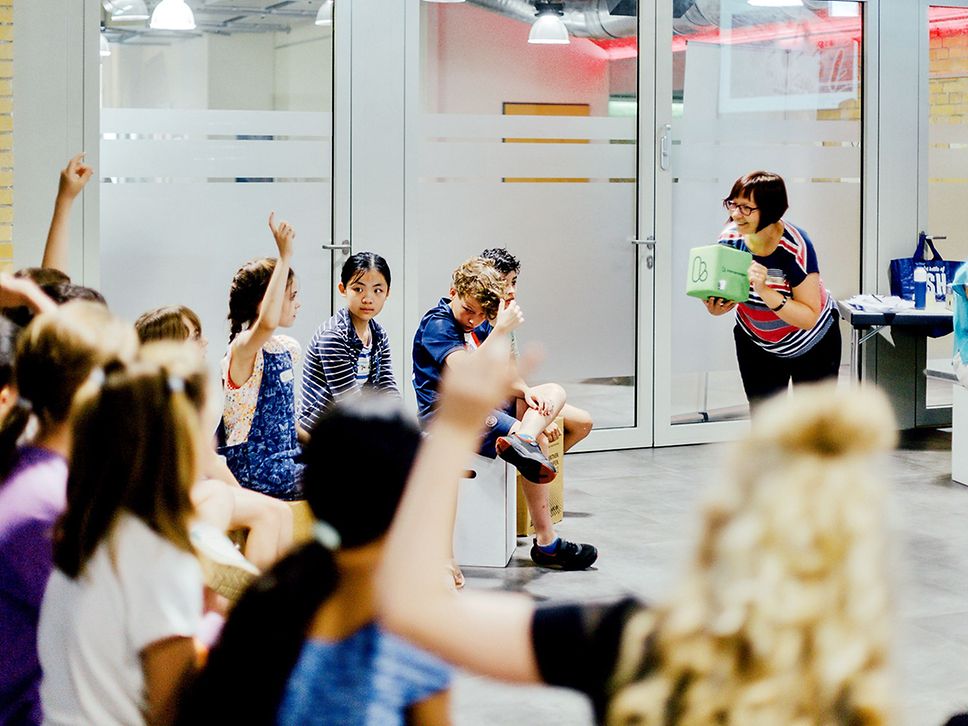 Jungs und Mädchen hören einer Frau zu, die einen grünen Würfel in der Hand hält. Ein paar Kinder melden sich.