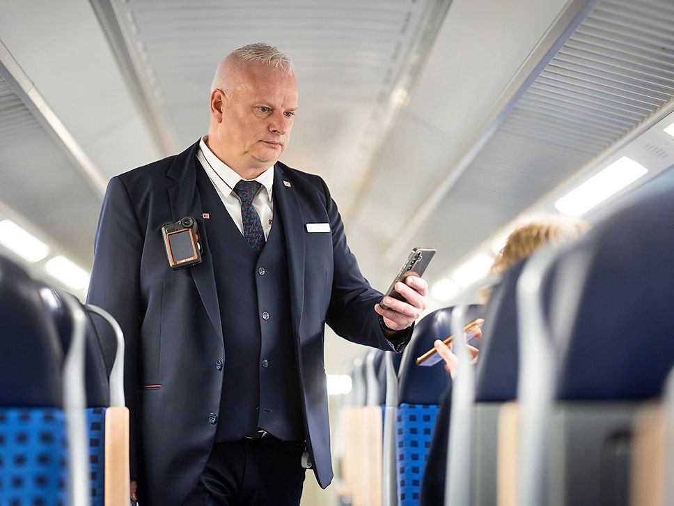 Ein Zugbegleiter kontrolliert Tickets in einem Regionalzug. Der Mann hat eine Bodycam am Revers.