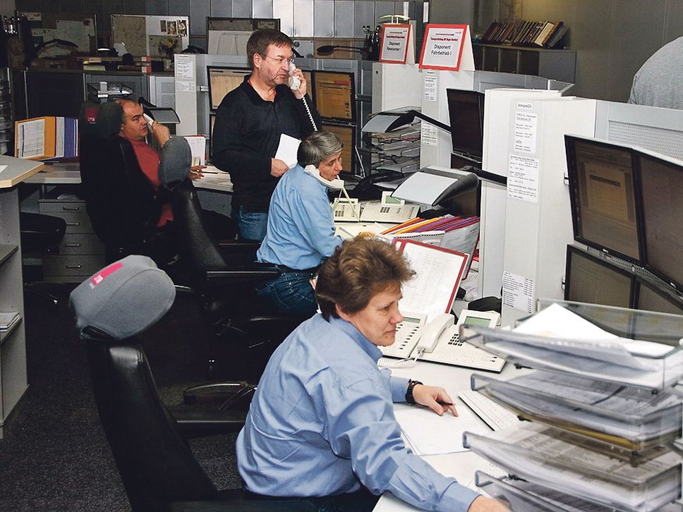Zwei Männer und zwei Sitzen vor Monitoren, drei der Personen telefonieren.