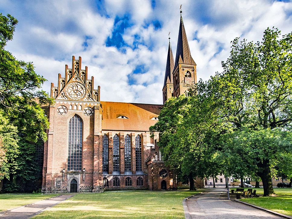 Dom St. Nikolaus in Stendal, ringsrum grüne Bäume. Der Himmel ist blau, aber stark bewölkt.