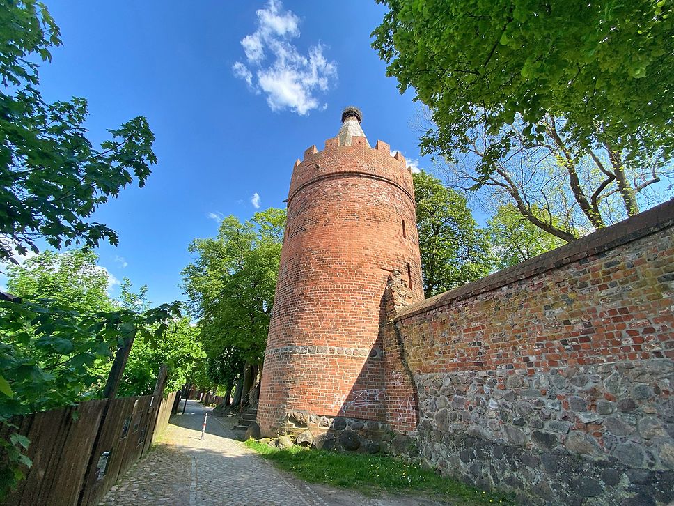Blick auf einen hohen Turm aus roten Backsteinen, rechts daneben eine Mauer.