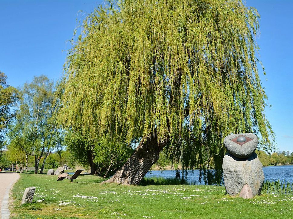 Skulpturen aus Stein stehen auf einer Wiese, im HIntergrund ein großer Baum, rechts ein See.