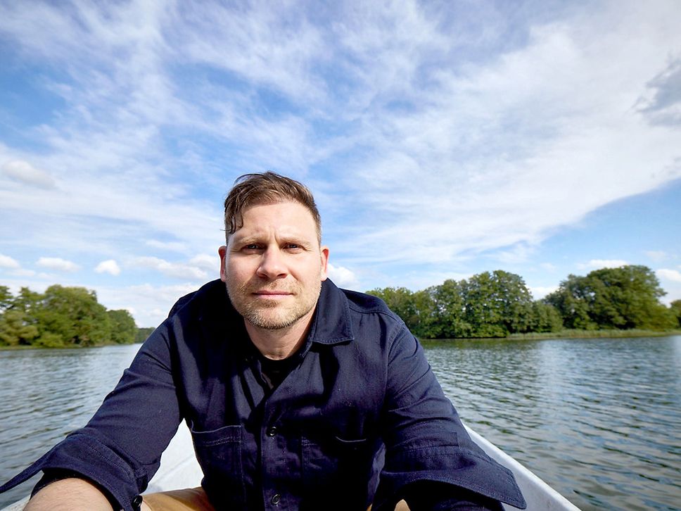 Ein Mann sitzt in einem Boot und schaut in die Kamera, im Hintergrund Wasser und Wald.