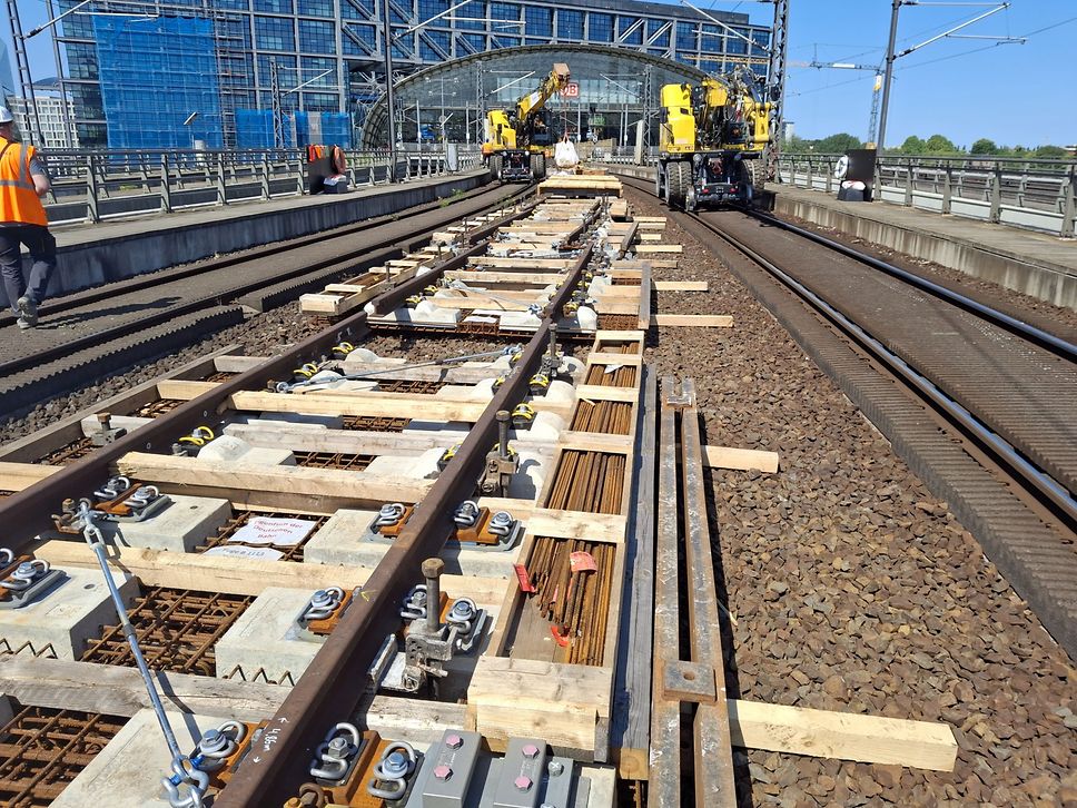 Man sieht, wie der Abschnitt einer Bahnstrecke erneuert wird, im Hintergrund Baumaschinen.