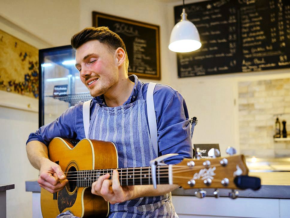 Ein Sänger mit blauem Hemd, Schürze und Gitarre spielt etwas. Er hat die Augen geschlossen.