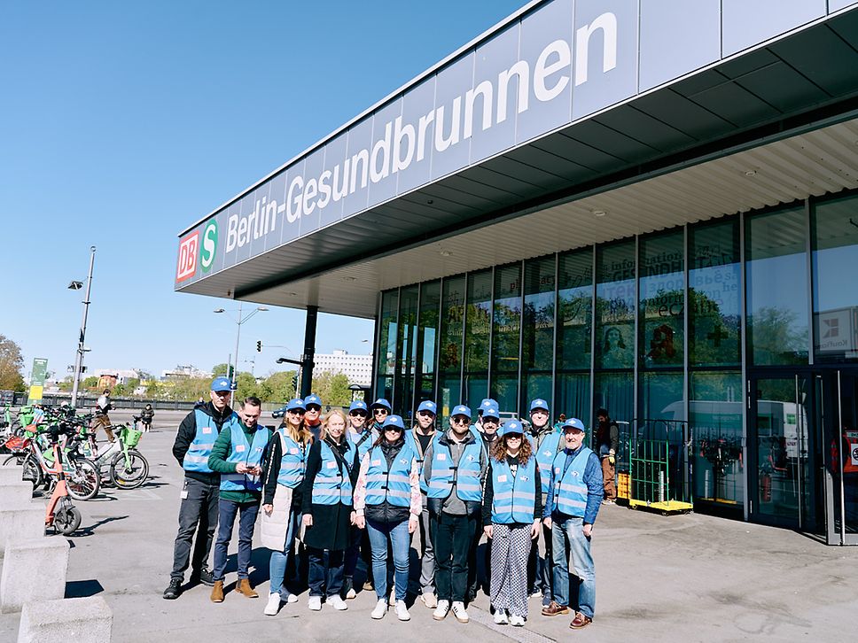 Eine Gruppe DB-Mitarbeitender in blauen Westen steht vor dem Bahnhof Berlin-Gesundbrunnen.