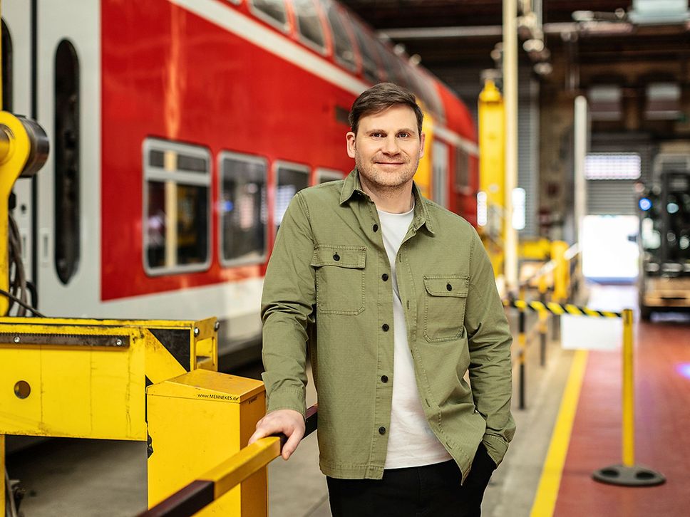 Ein junger Mann in grüner Jacke und weißem T-Shirt steht in einer Werkstatt vor einem roten Zug.