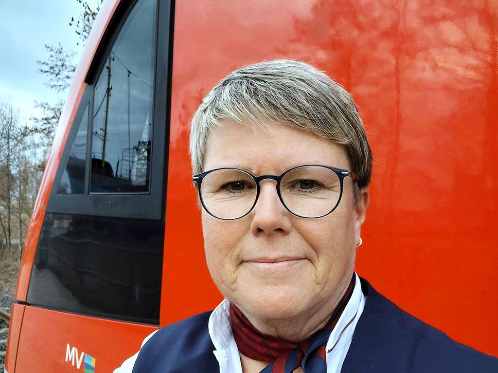 Selfie einer Frau mit kurzen blonden Haaren und schwarzer Brille. Die Frau steht vor einem roten Regionalzug.