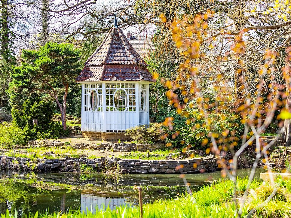 Pavillion im Park zwischen grünen Büschen und Bäumen, im Vordergrund Wasser.