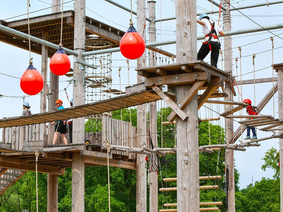 Vier Personen mit Klettergurten in einem mehrstöckigen Hochseilgarten aus Holz, Metallrohren und Seilen.