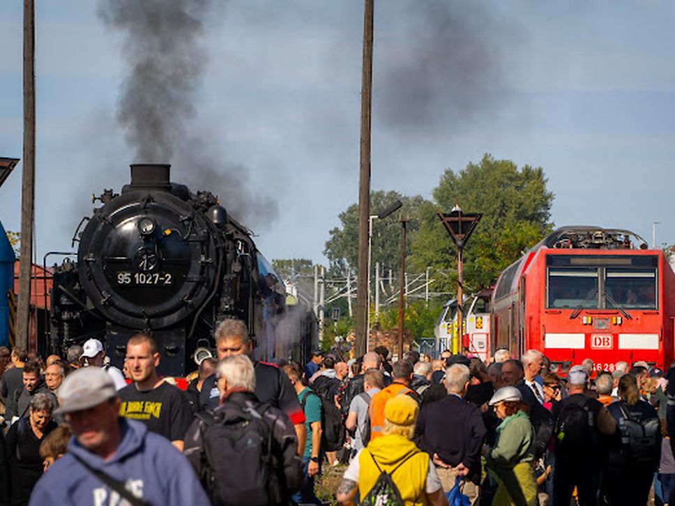Menschengruppe steht vor einer dampfenden, schwarzen Lok, rechts eine rote DB-Lok.