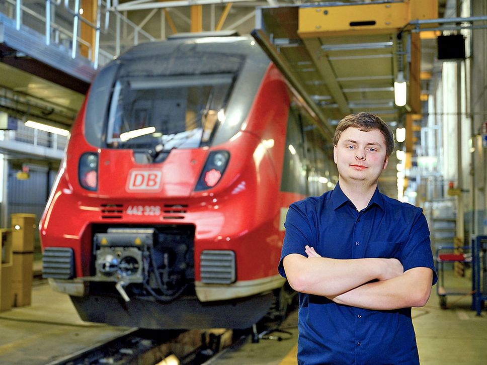 Portrait eines jungen Mannes, der mit verschränkten Armen vor einem roten Regionalzug steht.