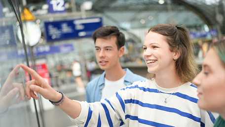 Drei Personen schauen lächelnd auf eine Informationstafel am Bahnhof