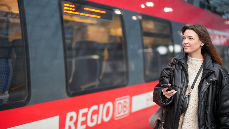 Eine junge Frau ist am Bahnhof mit Handy unterwegs und schaut lachend in die Umgebung.