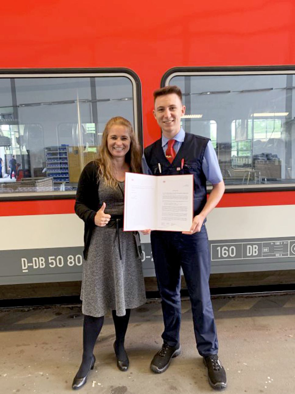 Eine Frau und ein Auszubildender in Uniform stehen mit einem Zeugnis vor einem roten Zug von DB Regio Nordost.