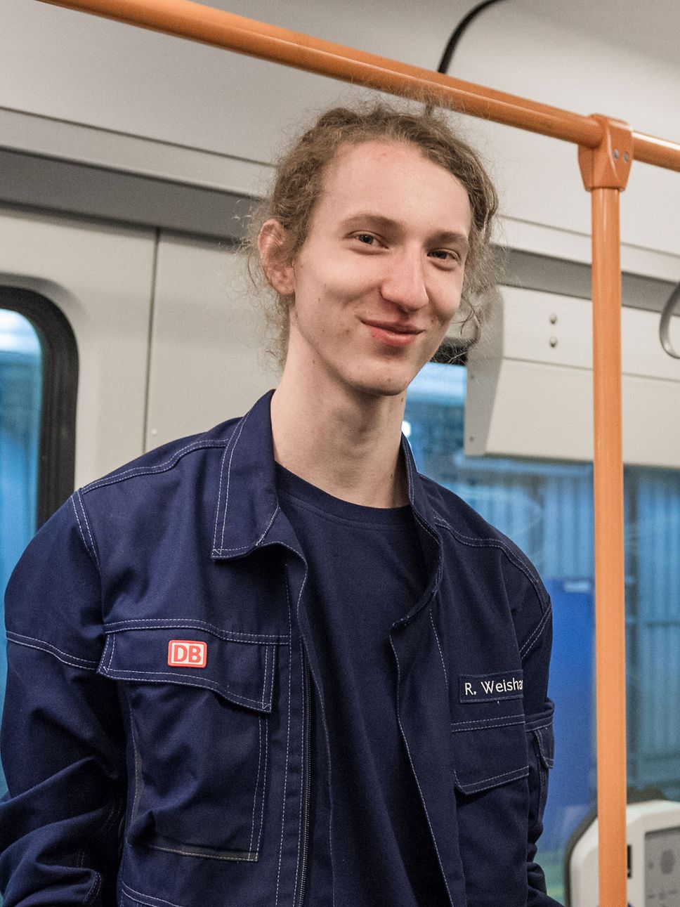 Ein junger Mann in Arbeitskleidung steht in einem Zug und lächelt in die Kamera.