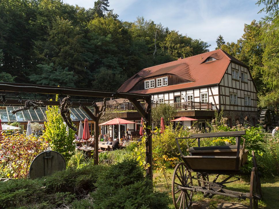 Fachwerkmühle mit Mühlrad und Restaurantterasse, im Vordergrund eine Kutsche
