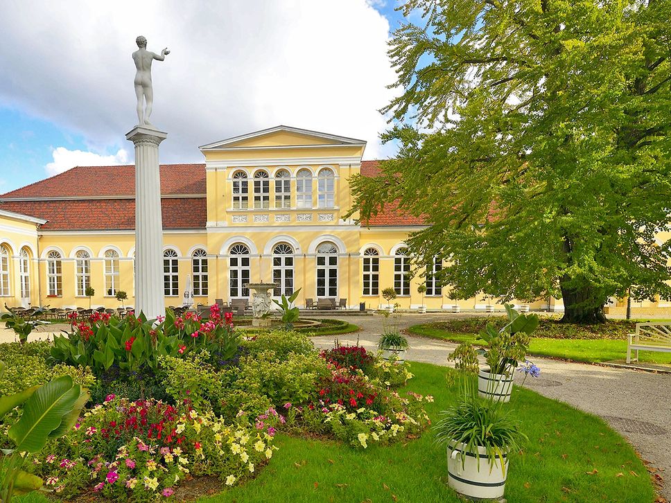 Die gelb gestrichene Orangerie mit Säulenfigur im Vordergrund.