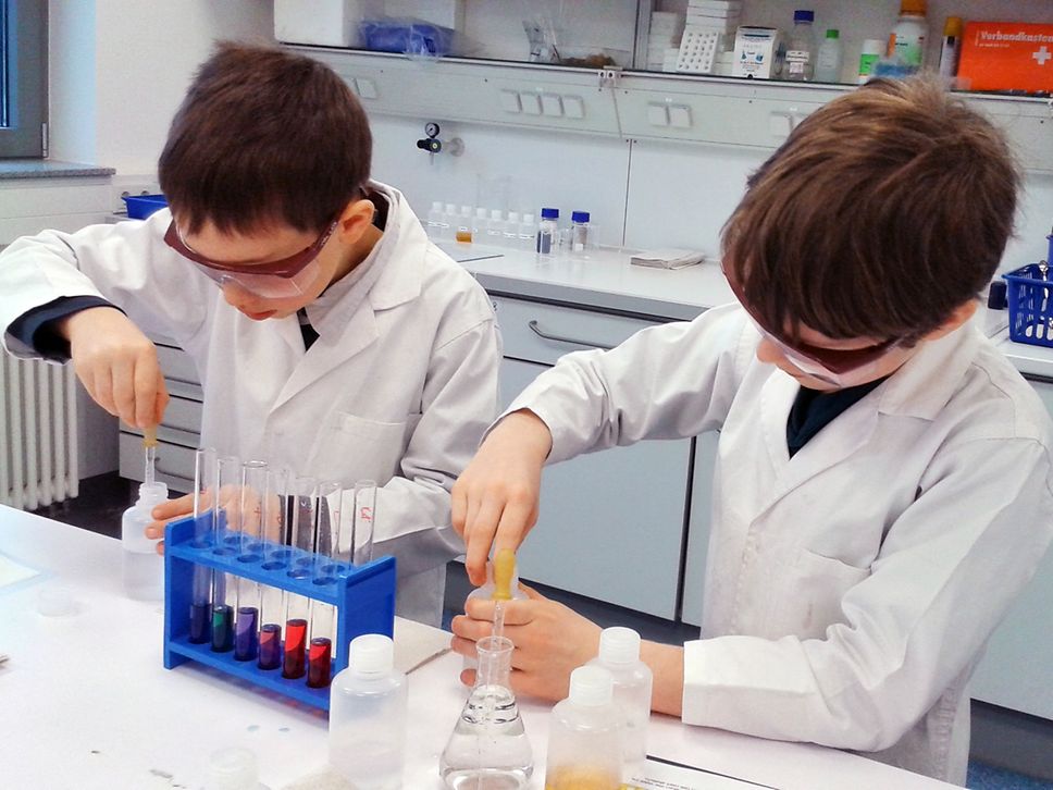 Zwei Jungs in weißen Kitteln und mit Schutzbrillen führen ein Experiment in einem Labor durch.