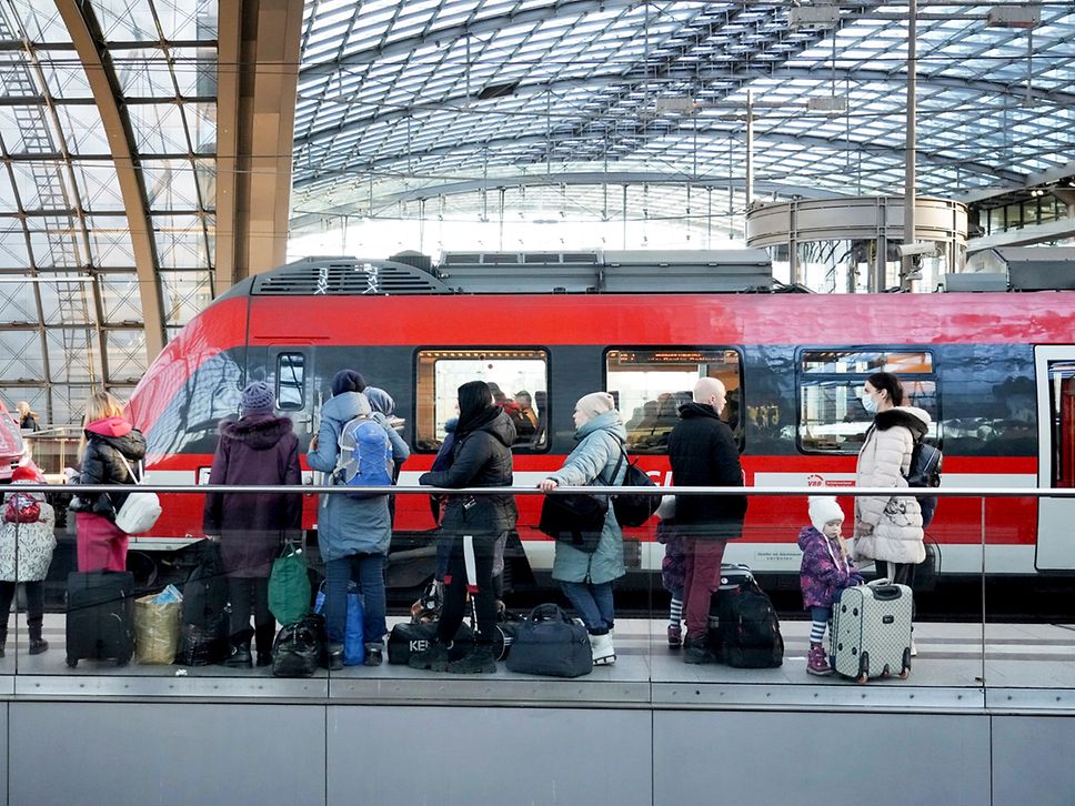 Menschen mit Koffern und Taschen stehen im Bahnhof vor einem roten Zug.