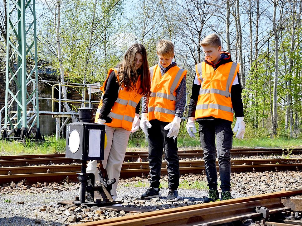 Ein Mädchen und zwei Jungs in orangfarbenen Warnwesten stehen zwischen Schienen an einer Weiche.