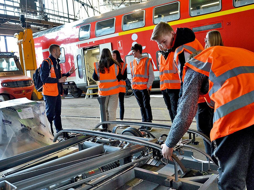 Schüler:innen in orangefarbenen Warnwesten stehen vor einem roten Zug und schauen sich Technik an.