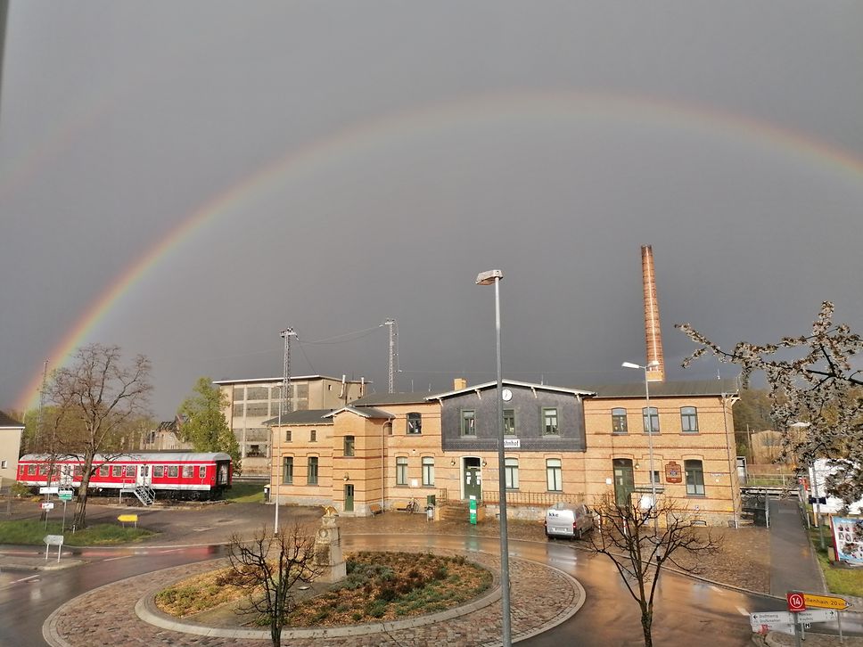 Ein Regenbogen vor grauem Himmel über dem Bahnhofsgebäude in Ortrand.