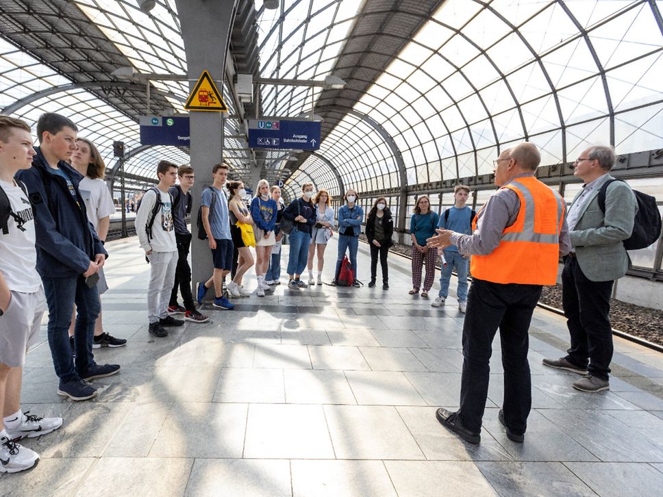 Eine Gruppe steht am Bahnsteig und hört einem Mann in Warnweste zu.