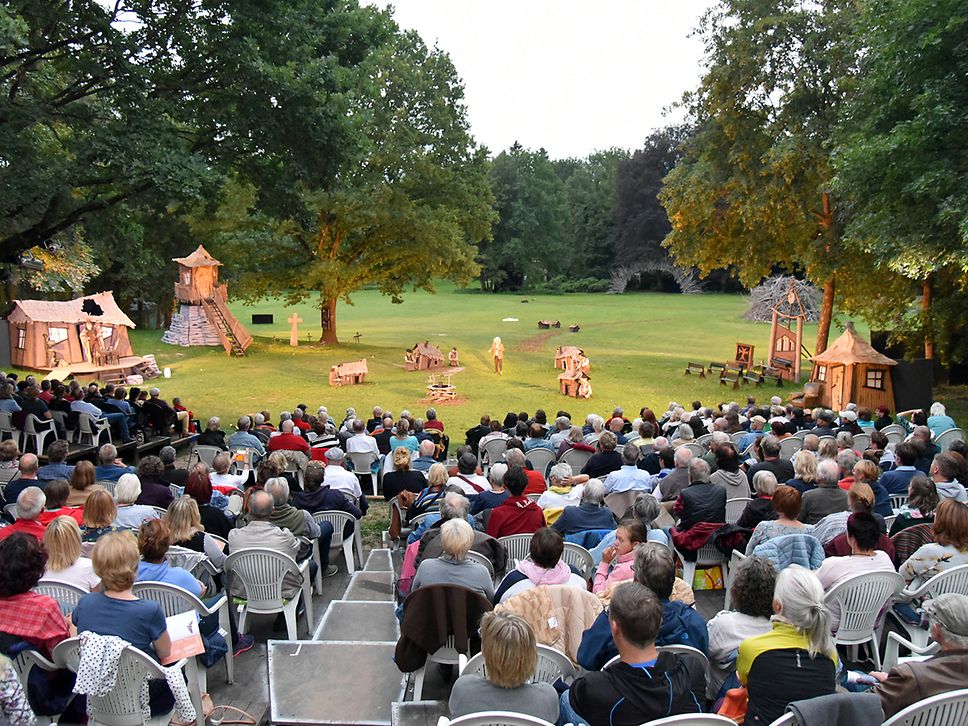 Publikum sitzt auf Stühlen und blickt auf Open-Air-Bühne, Wiese und Bäume