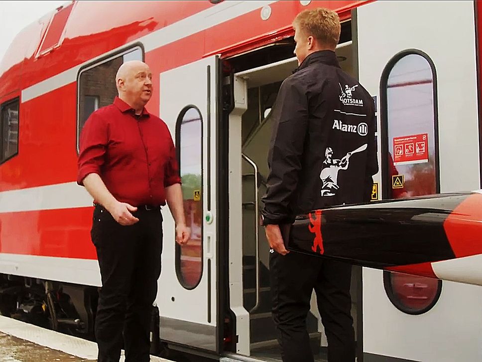 Kanute und Horst Evers stehen vor einem roten Zug und der Kanute will mit Kanu ins Fahrzeug.