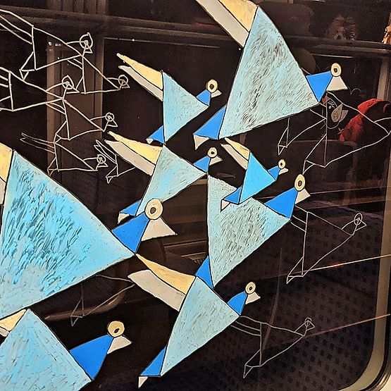 Fensterbild am Innenfenster eines Zuges, zu sehen sind blaue Vögel in abstrakter Form mit Körpern aus Dreiecken.