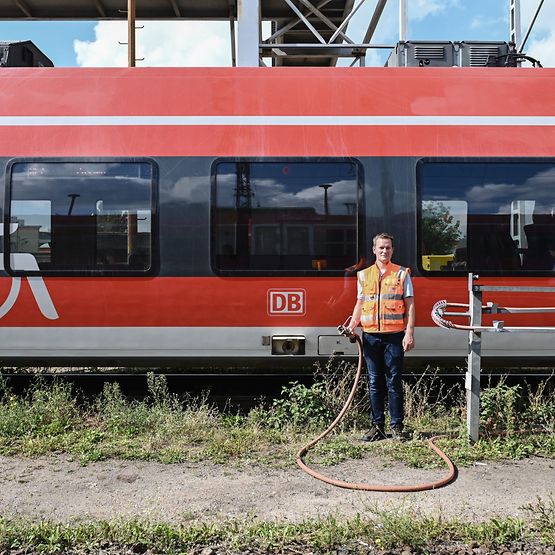 Ein Mann in orangefarbener Warnweste steht mit Schlauch in der Hand vor einem roten Zug.