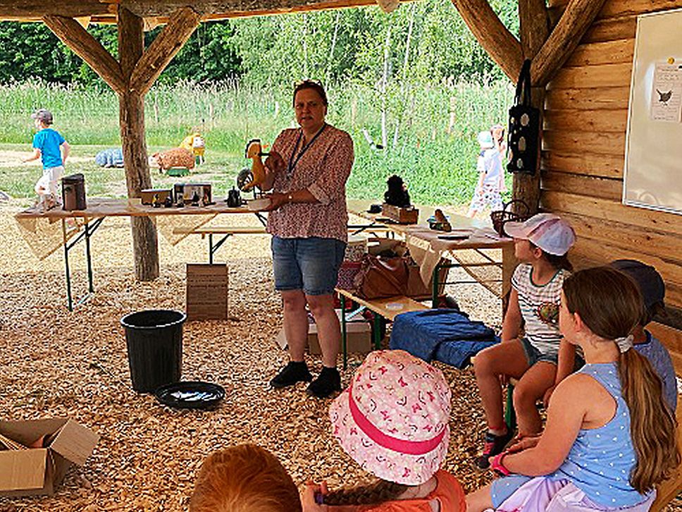 Kinder sitzen auf Bänken und lassen sich von einer Frau etwas erklären.