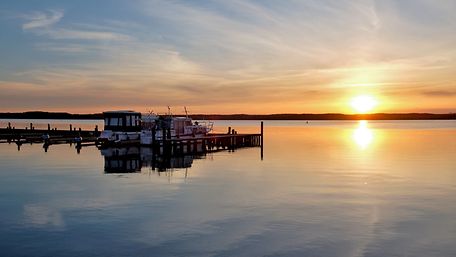 Sonnenuntergang an einem See, links im Bild ein Steg mit Booten.