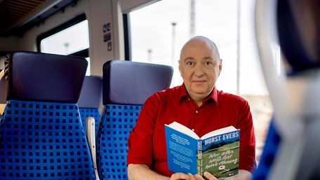 Kabarettist und Autor Horst Evers sitzt in einem Regionalzug und hat eines seiner Bücher aufgeschlagen in der Hand.