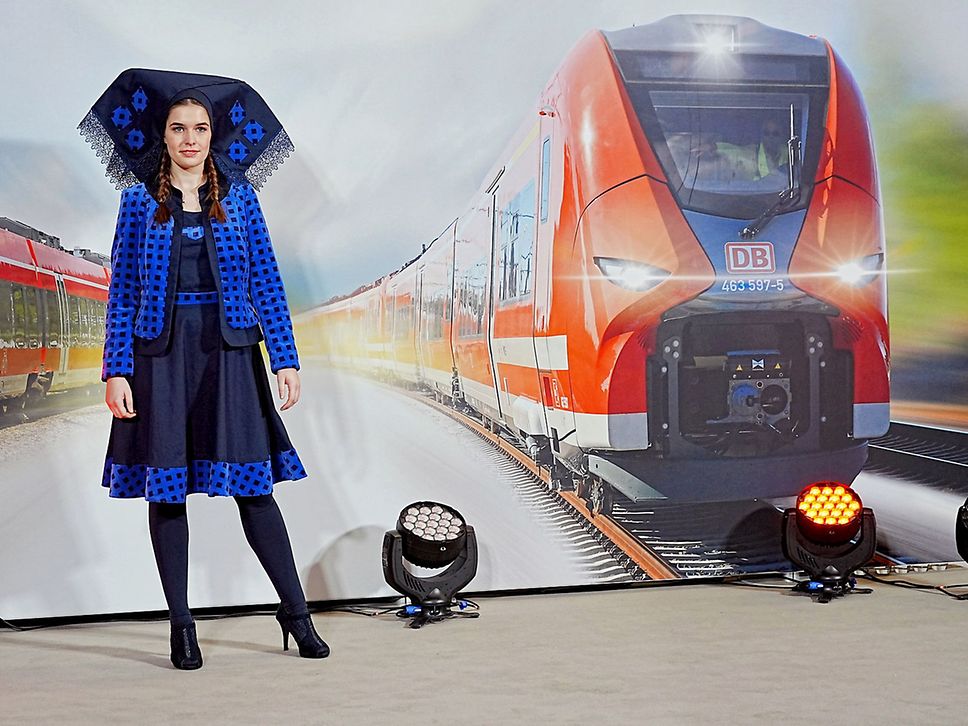 Eine Frau in einem blauen Designer-Outfit steht vor einer Wand, auf der ein roter Regionalzug zu sehen ist.