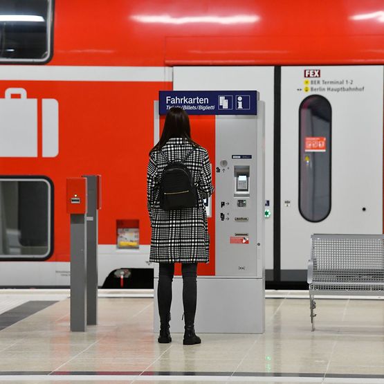 Eine Frau ist von hinten zu sehen, sie steht an einem Fahrausweisautomaten, im Hintergurnd eine rote Regionalbahn.